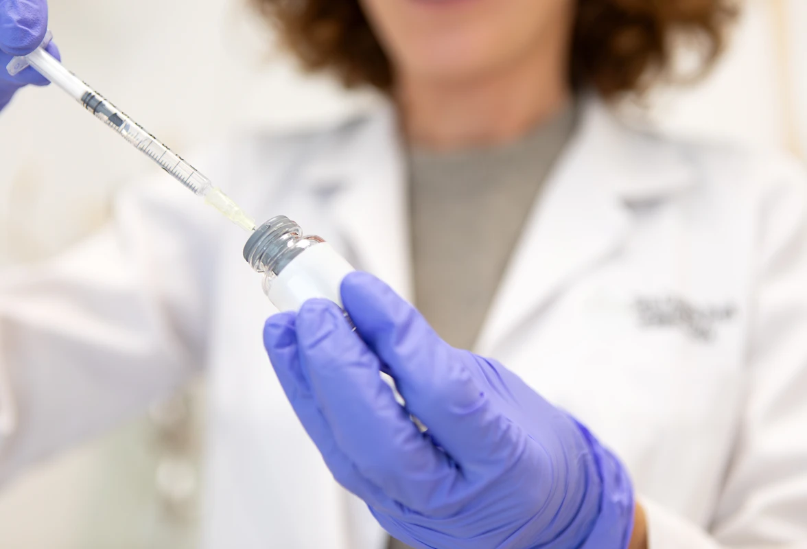 Woman in a white lab coat and blue gloves holding a syringe, likely administering a vaccine to a patient